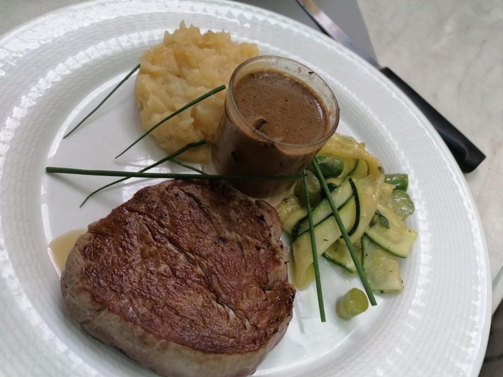 Un pavé de bœuf avec une purée, des légumes verts et un petit pot de sauce